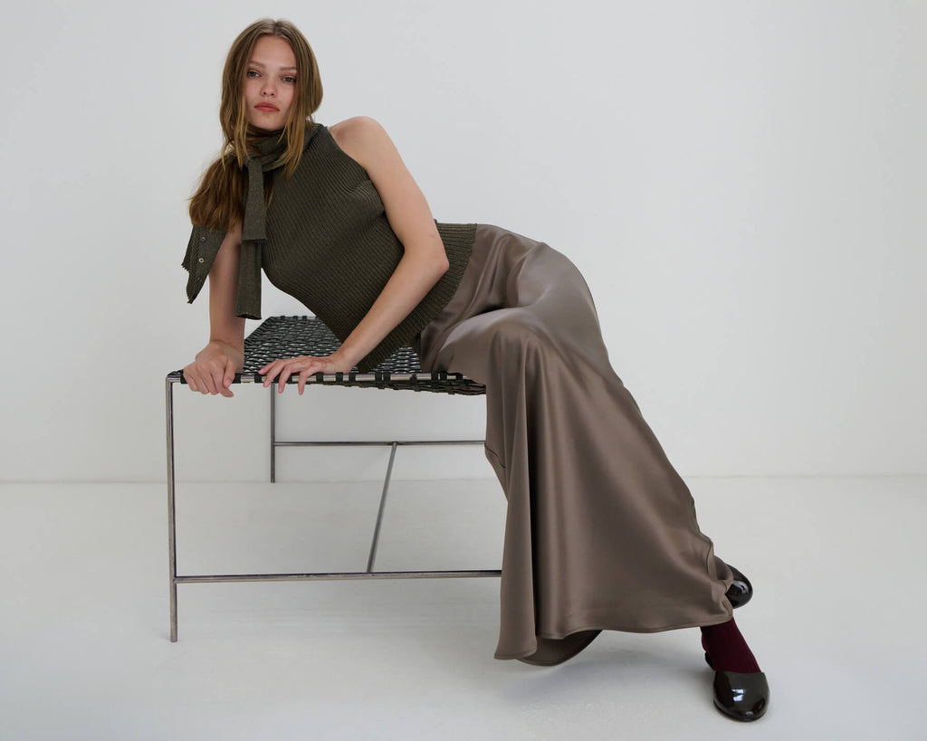 Woman leaning on stool in taupe Barb satin maxi skirt, mules and socks, and Blake moss cardigan and sleeveless mockneck.