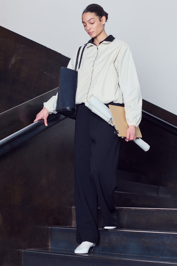 Woman walking down stairs in Kyoto windbreaker zipped up, with white heels and bag, holding a clipboard.