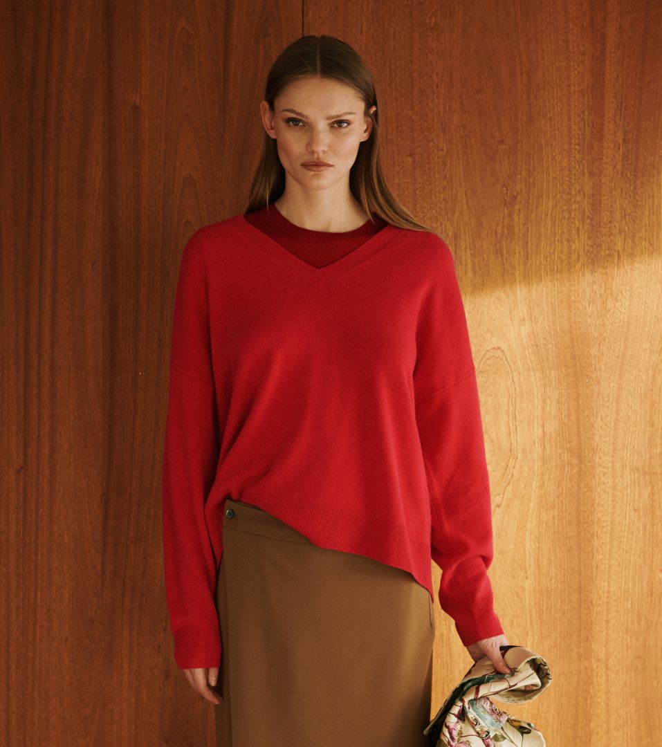 Model is standing in front of wooden wall in red v neck sweater and brown camel skirt.