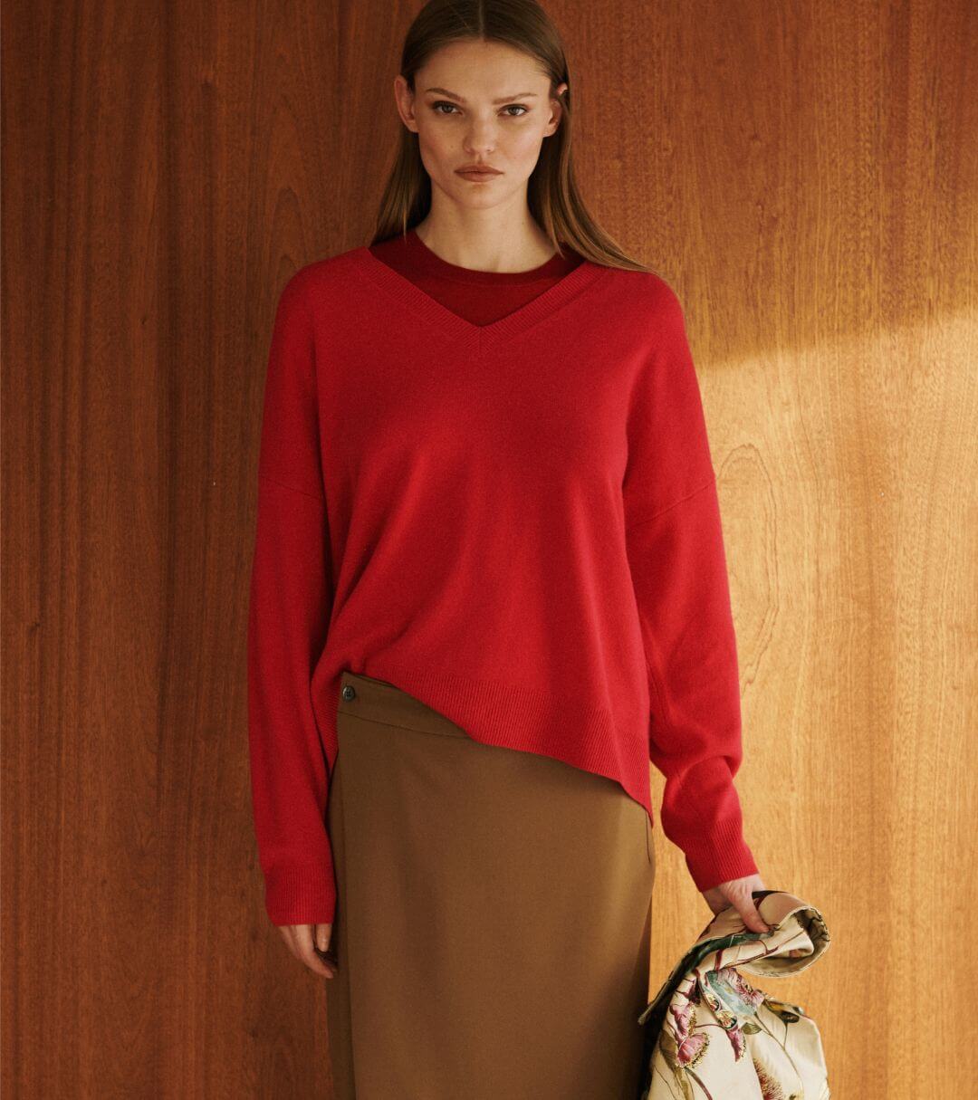 Model is standing in front of wooden wall in red v neck sweater and brown camel skirt.