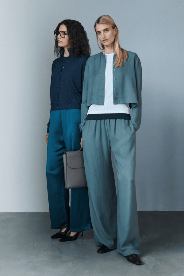 Two women in cardigans and trousers standing with hands in pockets, one with briefcase.
