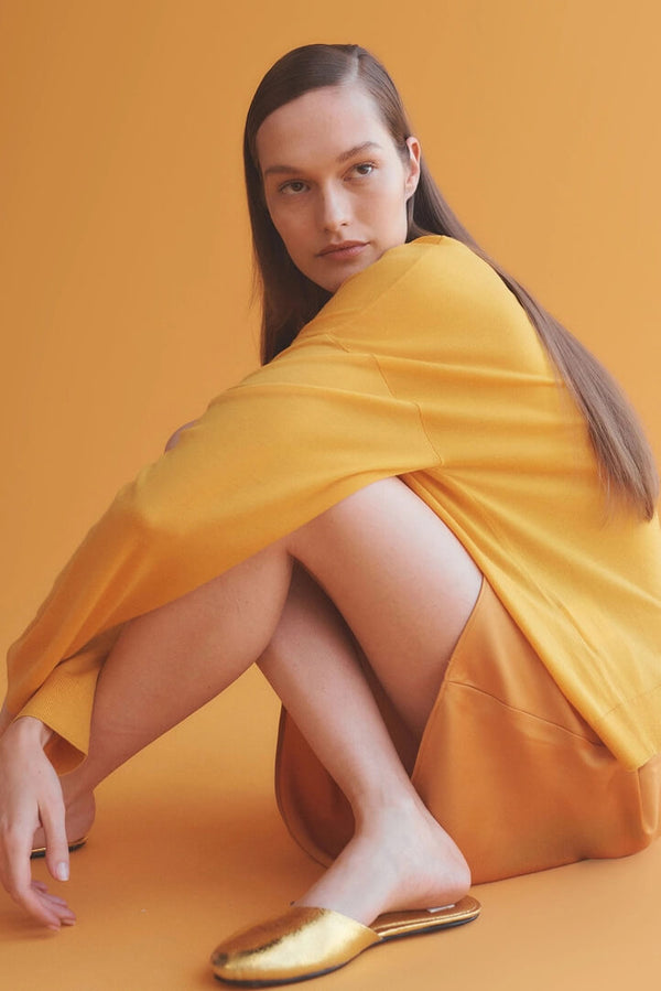 Woman sitting in orange sweater and slip skirt with slides, with orange background.