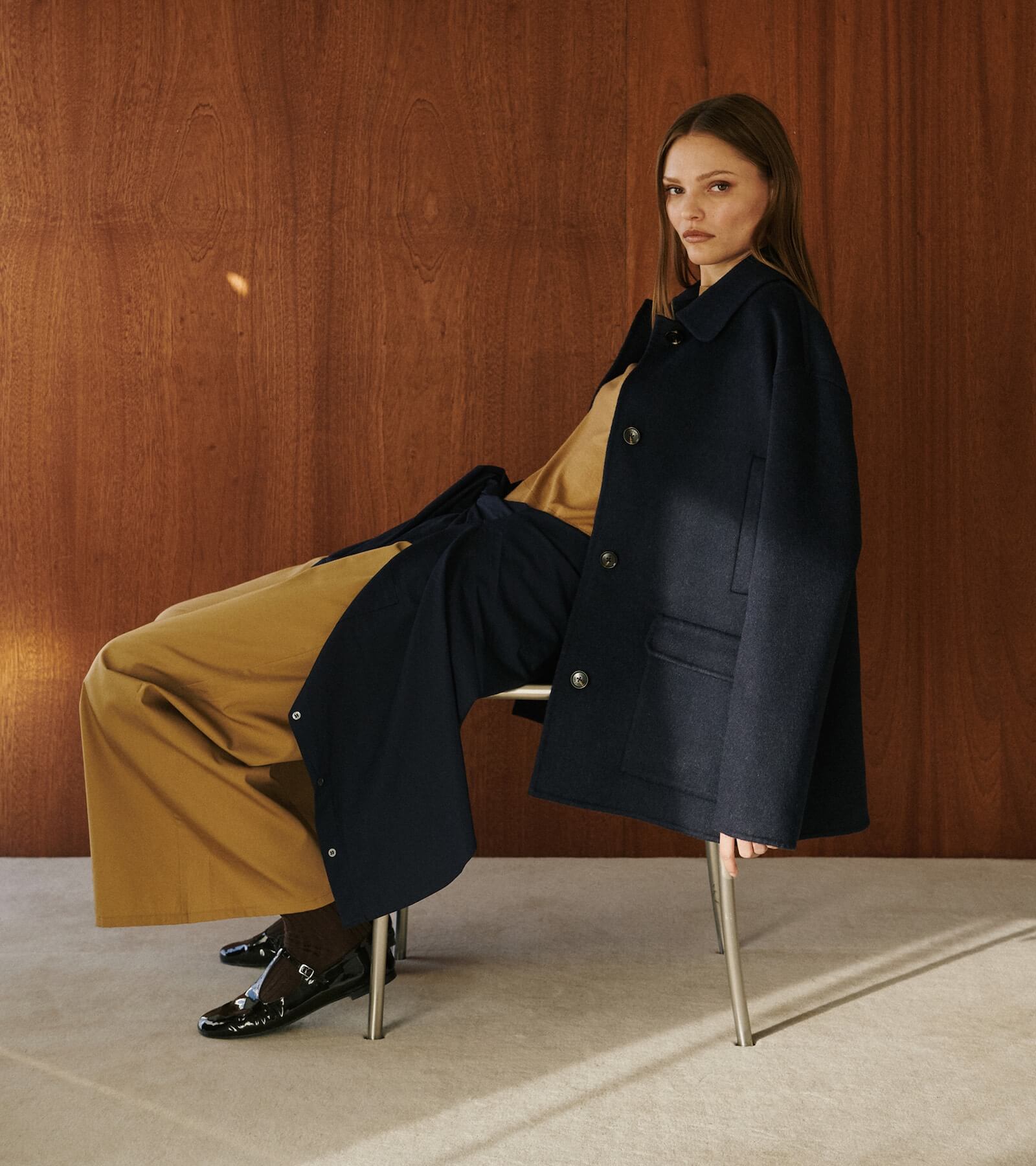 Woman sitting in chair in front of wooden wall in camel colored pants and top with coat draped over.