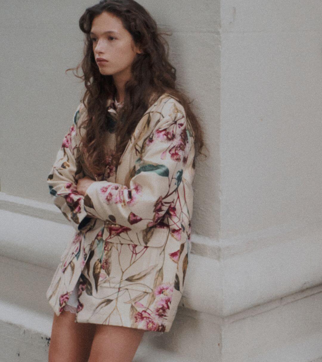 Model is standing in floral coat with arms crossed, against wall
