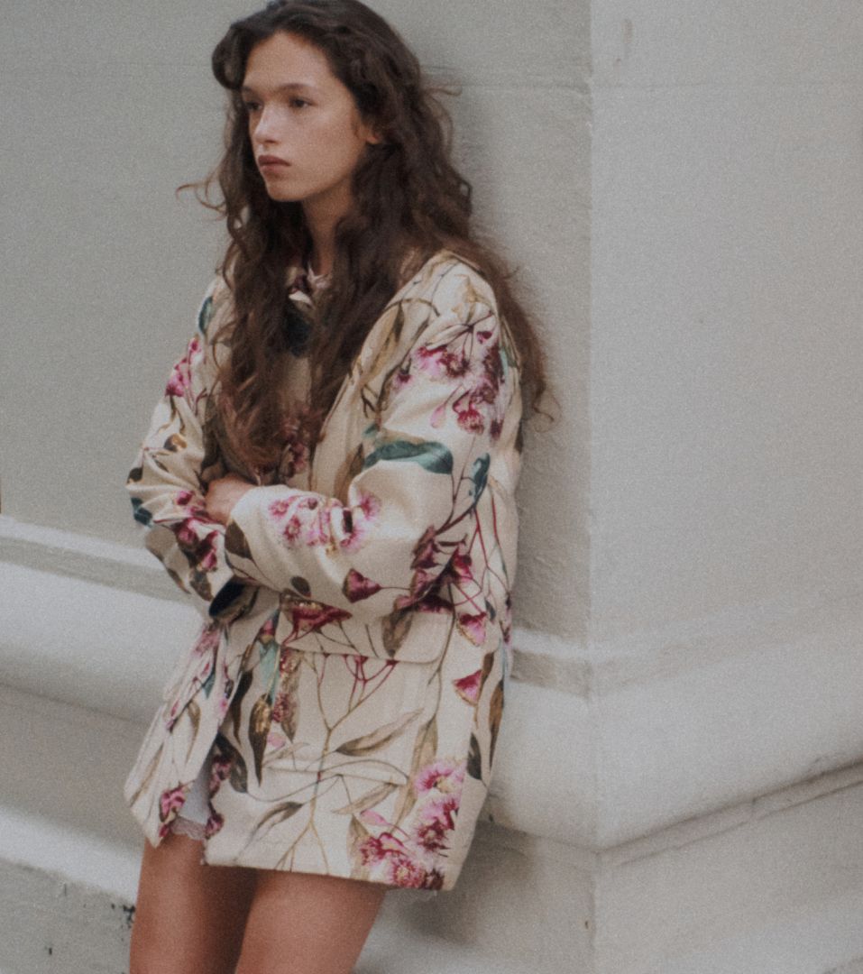 Model is standing in floral coat with arms crossed, against wall