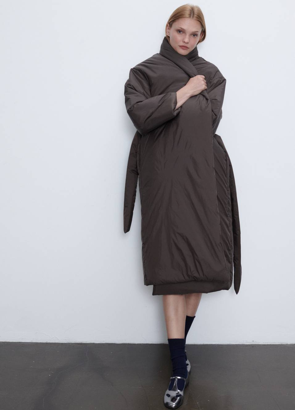 Model is standing in bo puffer coat in brown with socks and black mary jane shoes.