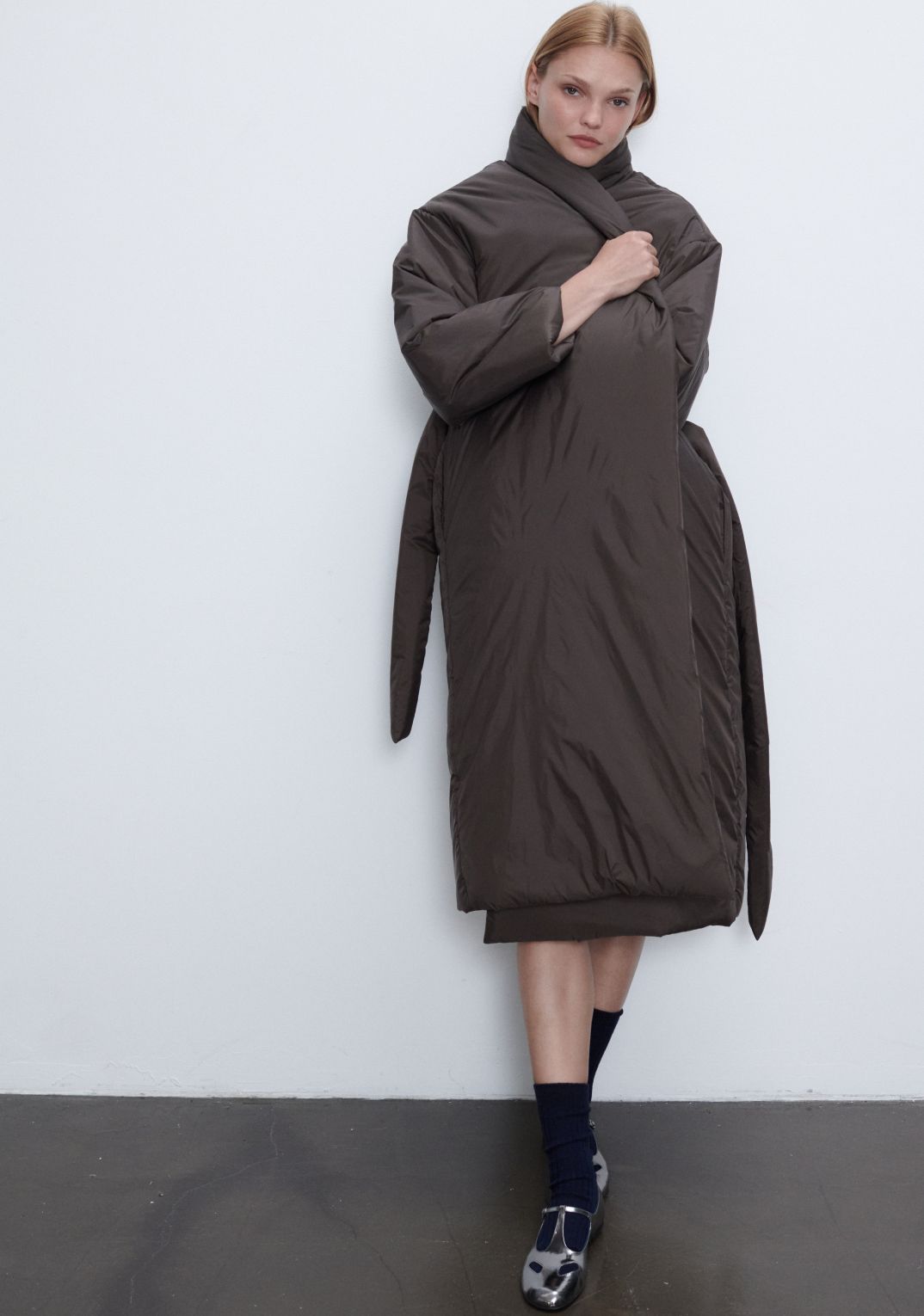 Model is standing in bo puffer coat in brown with socks and black mary jane shoes.