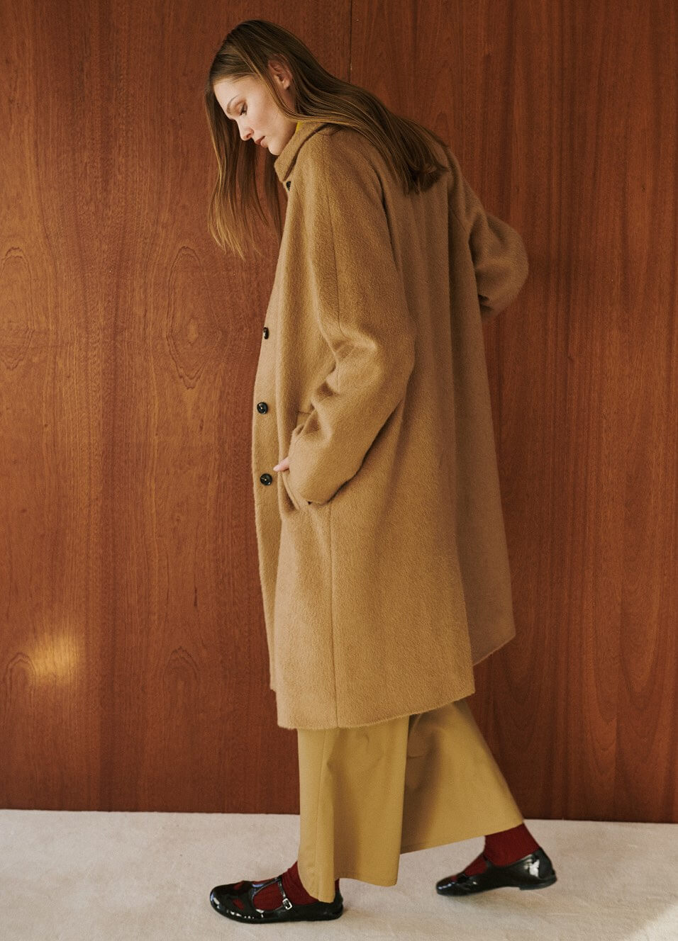 Close up of woman wearing long camel coat and beige pants, in front of wooden wall
