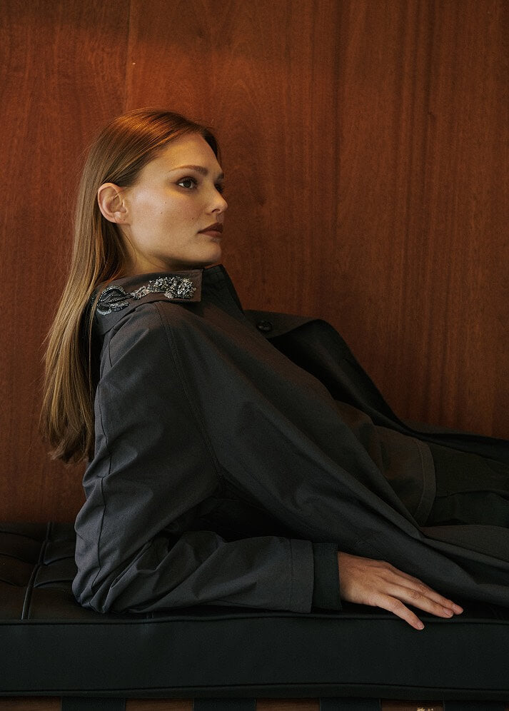 Model is leaning down wearing grey coat with embroidery detail.