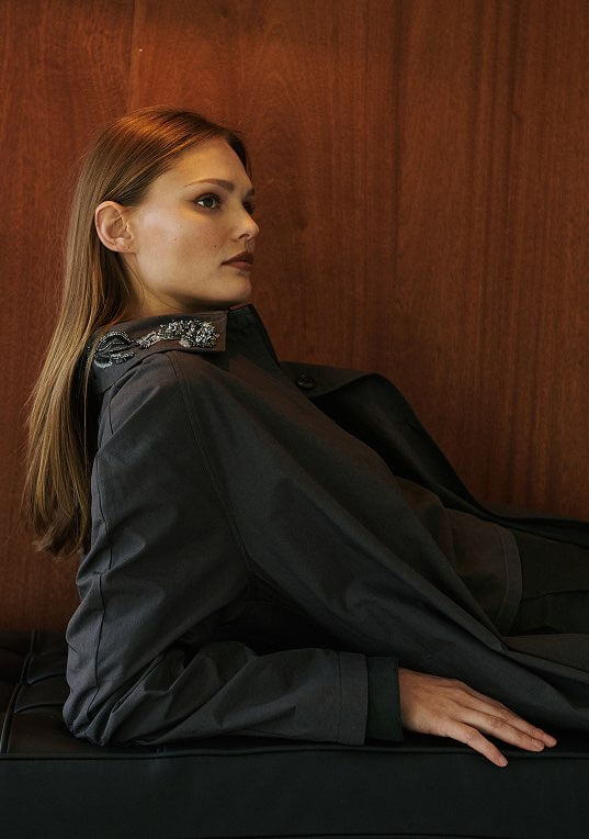 Model is leaning down wearing grey coat with embroidery detail.