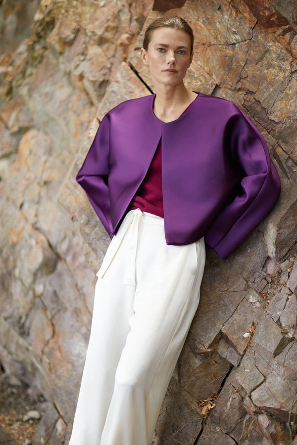 Woman leaning again outdoor wall in Bobbi purple jacket and Barb creme tailored pant