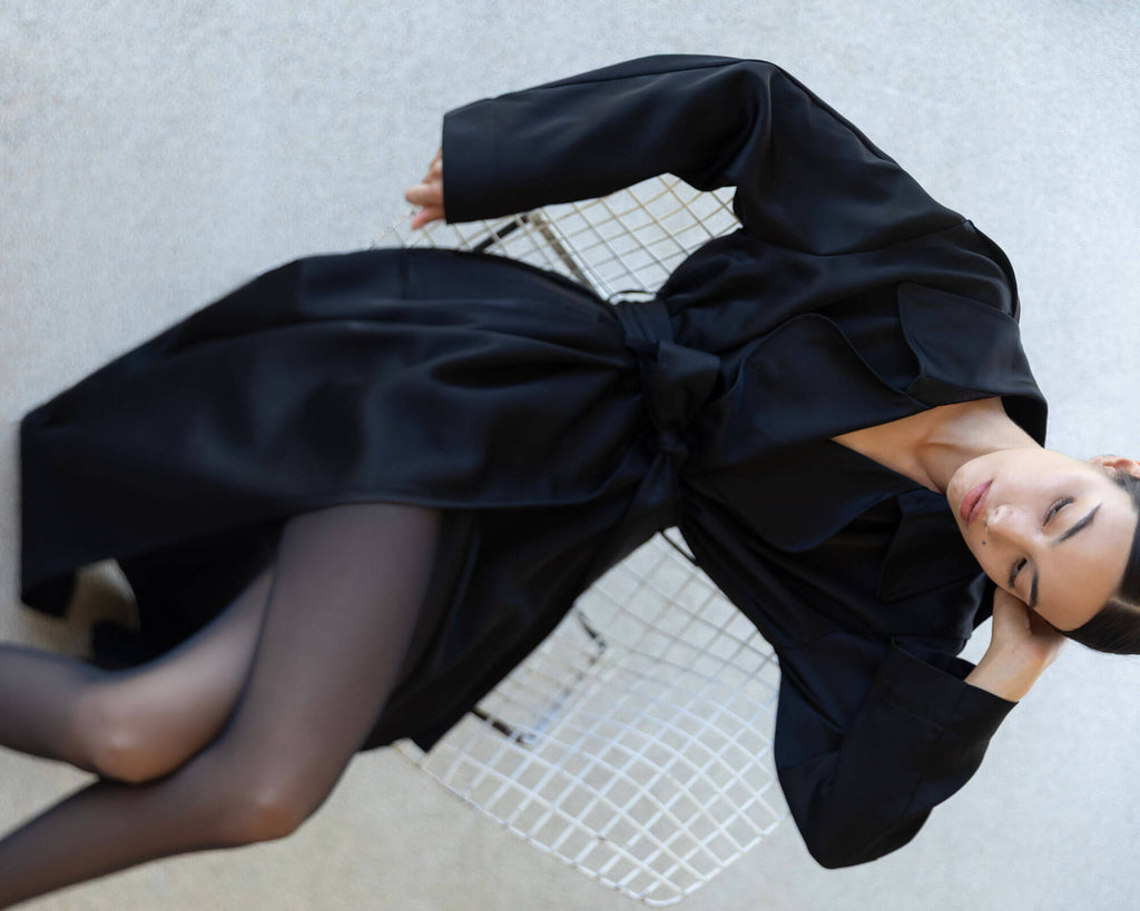 Woman leaning on chair in barb black satin trench and tights, looking to the side