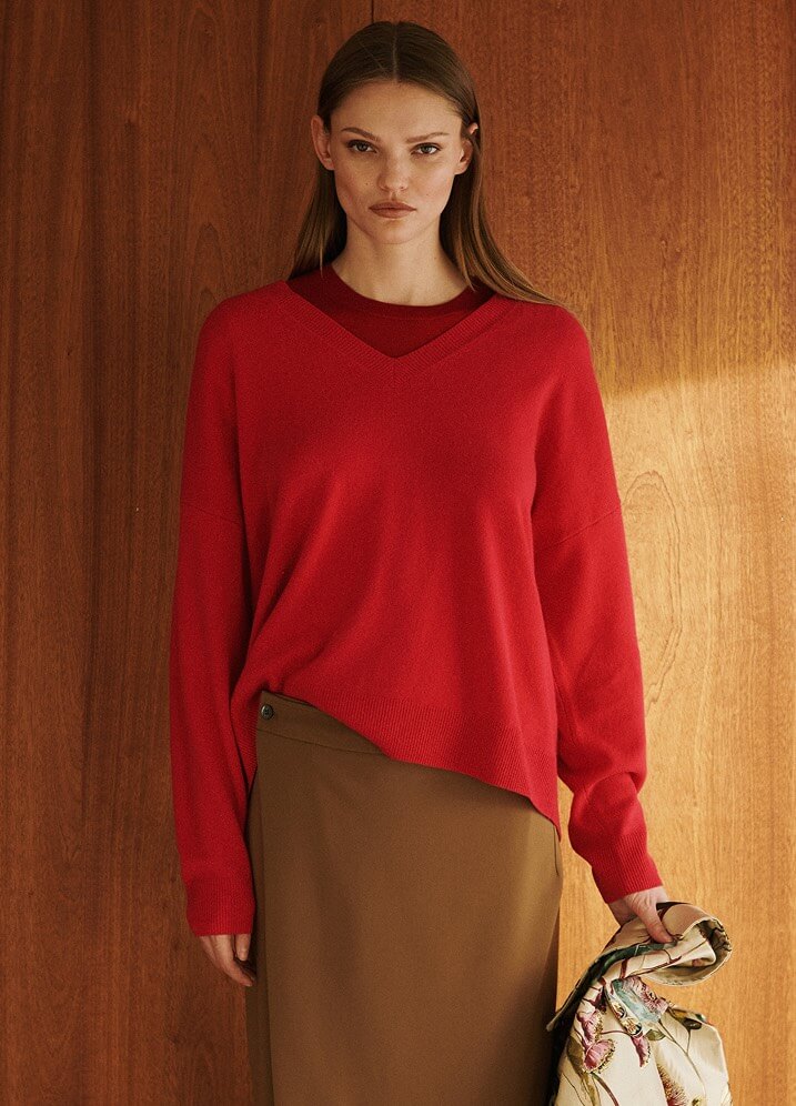 Model is wearing red v neck sweater and brown skirt standing in front of wood wall