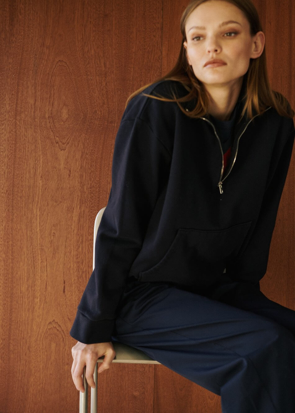 Woman sitting in chair in navy anorak in front of wooden wall.
