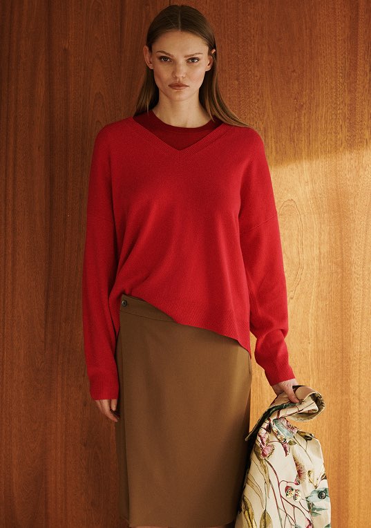 Model is wearing red v neck sweater and brown skirt standing in front of wood wall