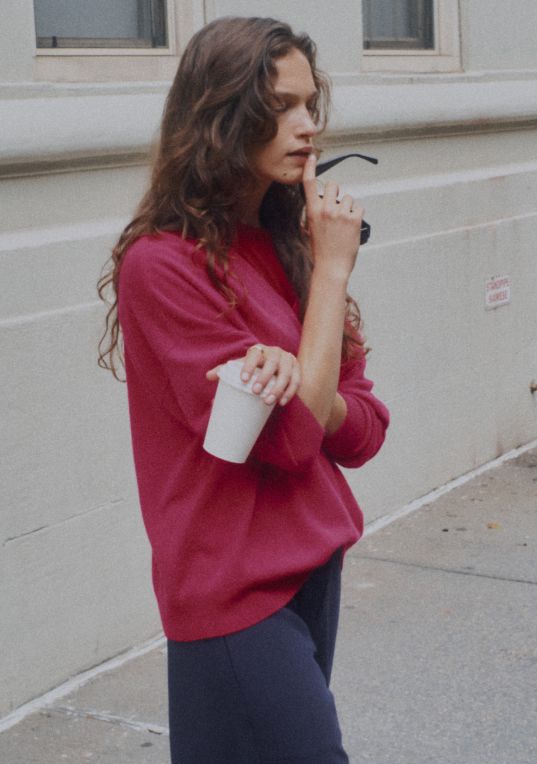 Model is standing holding cup and sunglasses in red sweater.