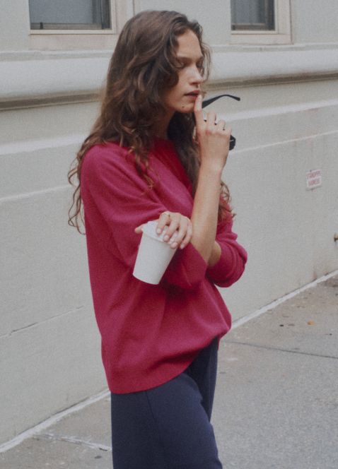 Model is standing holding cup and sunglasses in red sweater.