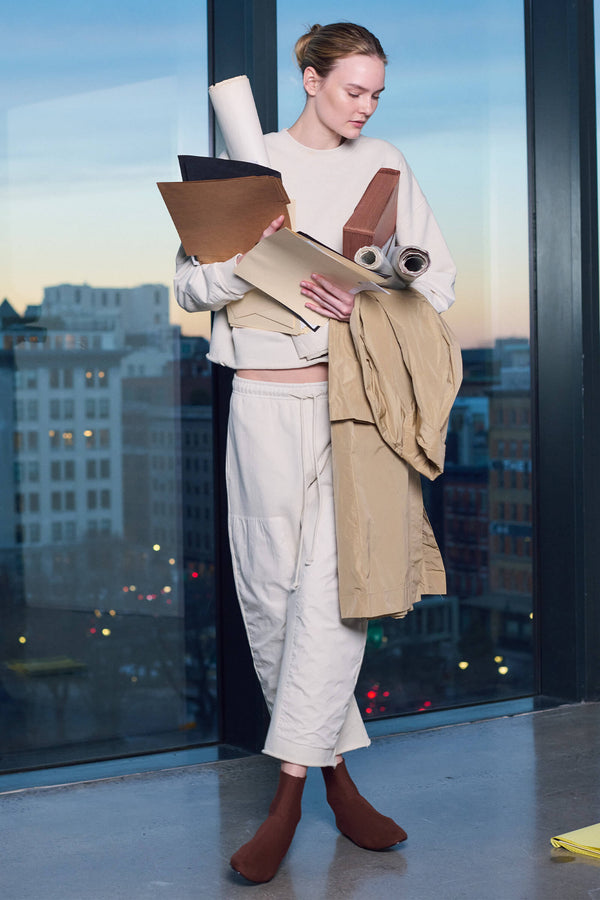 Woman standing in front of window with a city view, holding notebooks, papers, and a coat, looking down at a notebook pad on the floor.