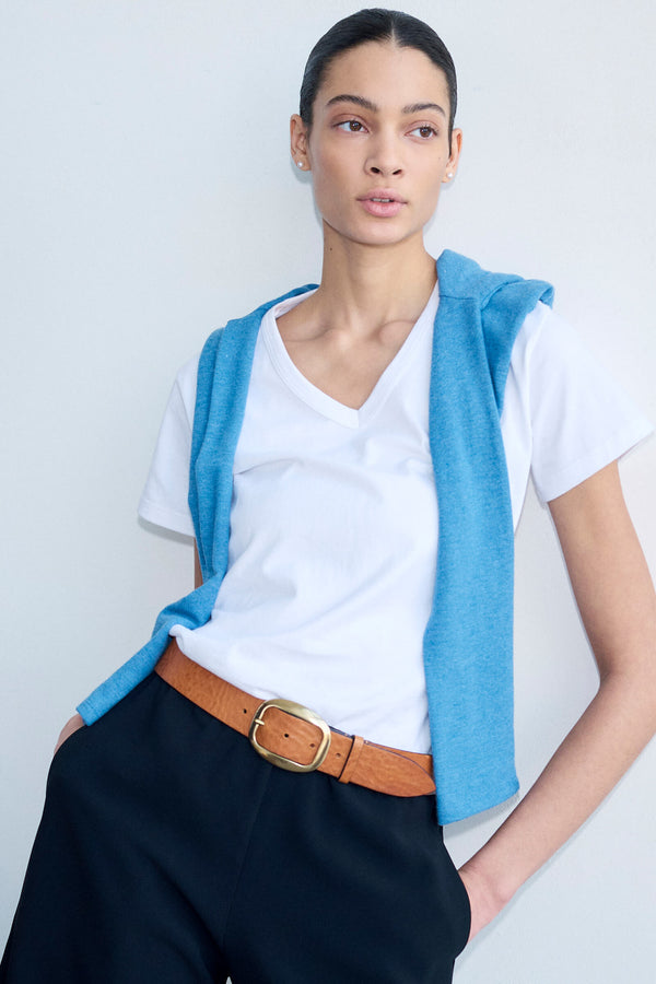 Woman standing in front of blank wall in white V neck tee, with blue sweater draped over and brown belt.