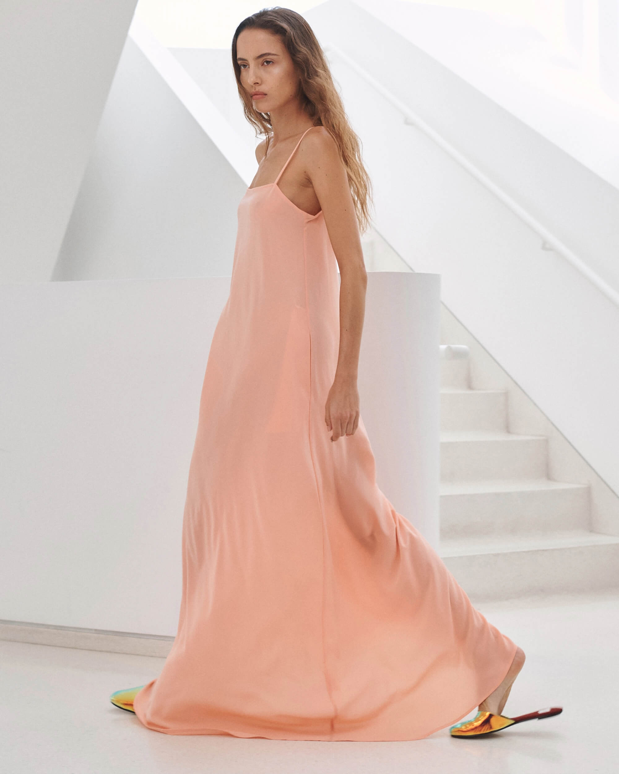 Woman in maxi peach dress with slides, in front of white wall and steps.