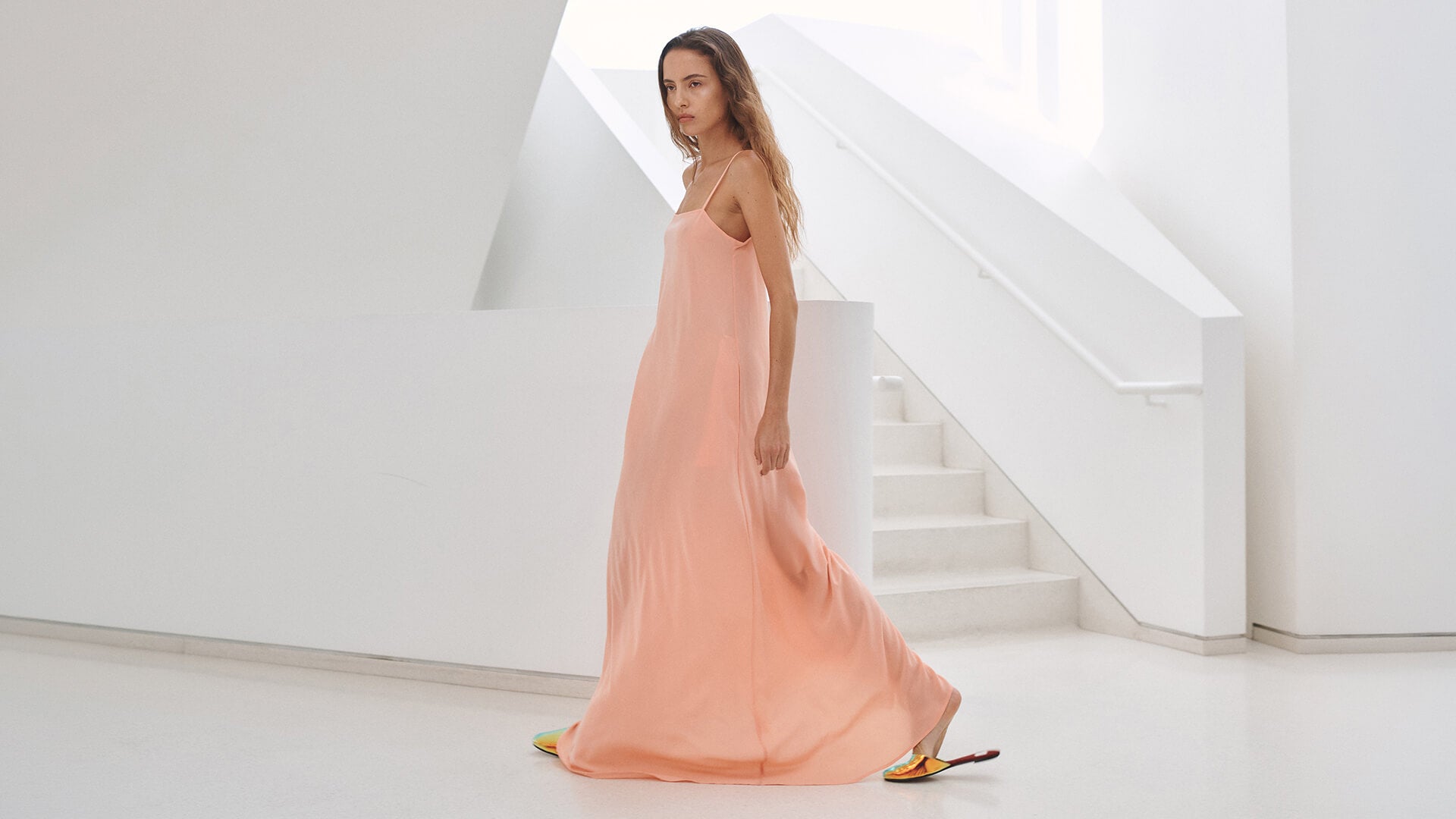 Woman in maxi peach dress with slides, in front of white wall and steps.
