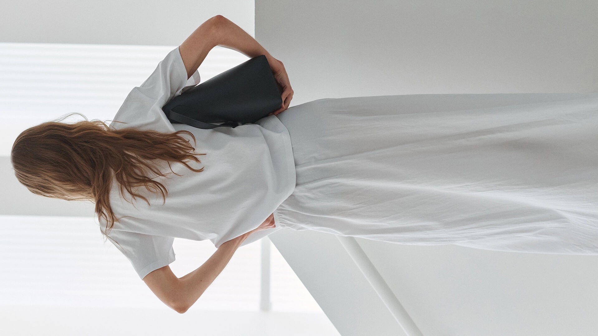 Back of woman in white t-shirt and maxi skirt, facing the wall, holding black purse.