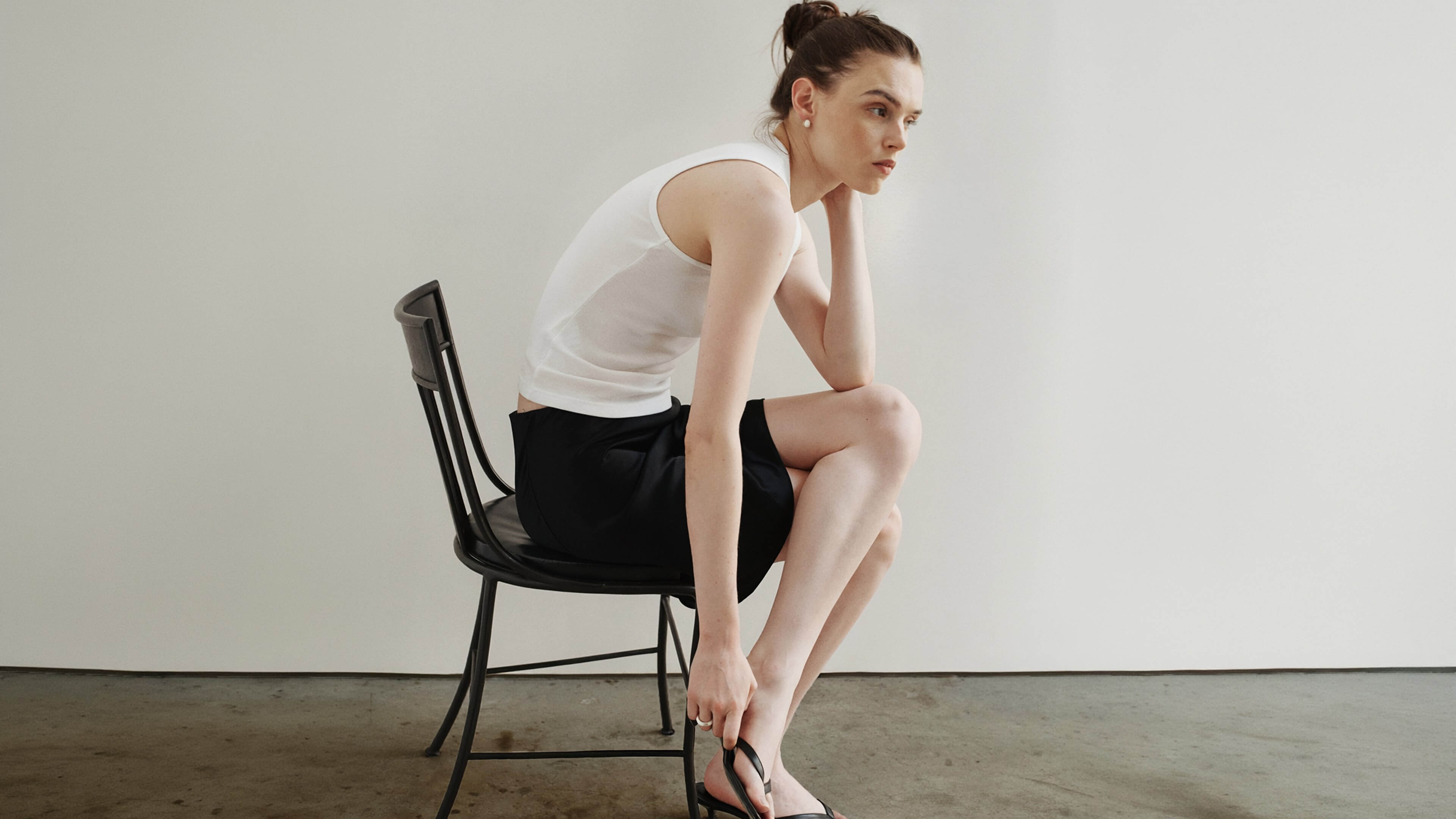 Woman sitting in chair in black skirt and white tank, hunched over.