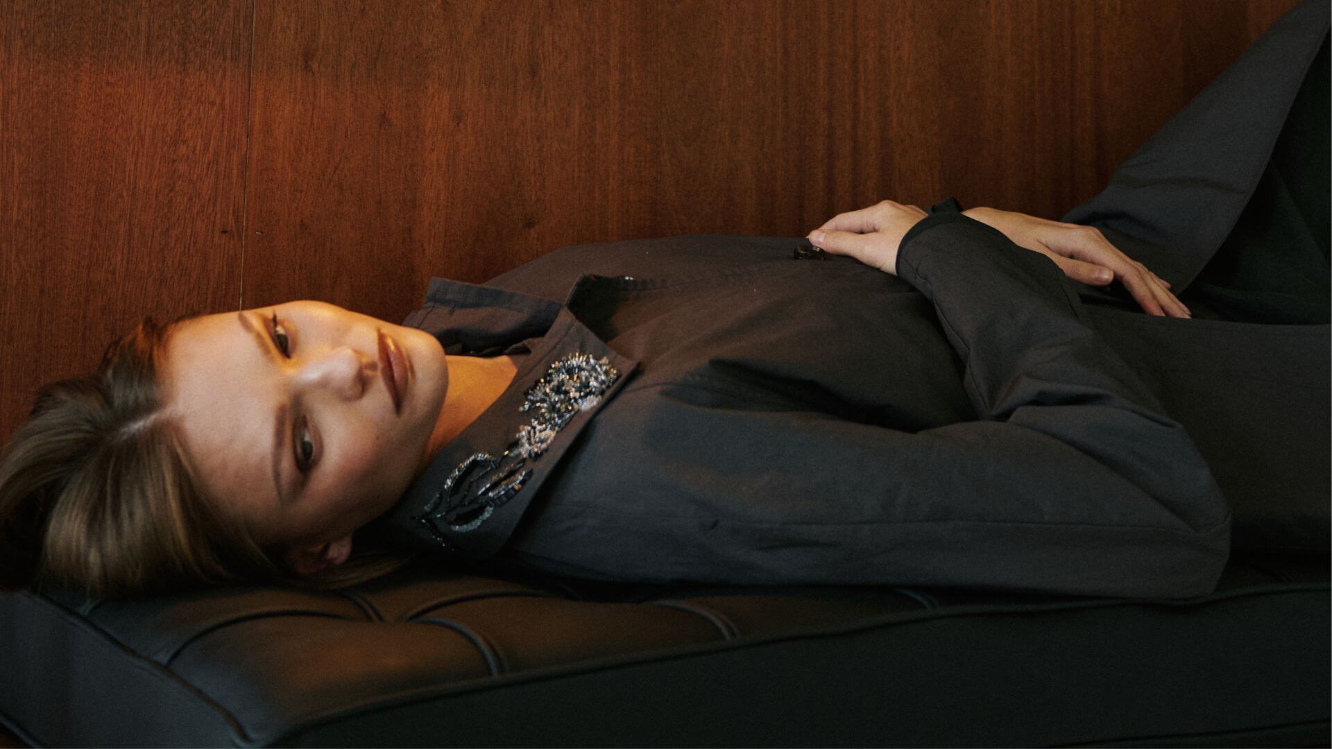 Model is laying down on couch in embroidered kyoto car coat in slate.