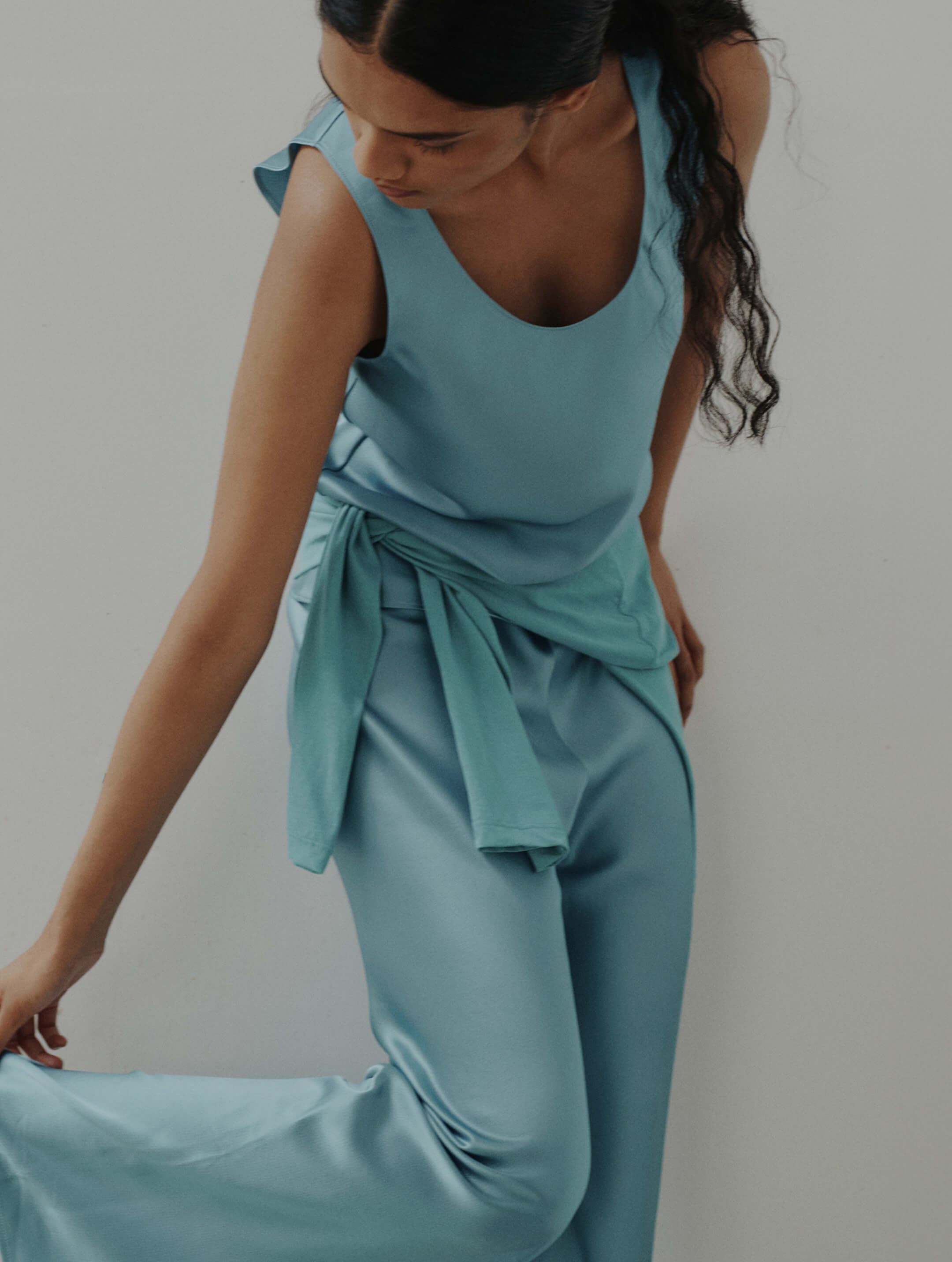 Side view of woman wearing blue satin tank and matching pants, holding ankle.